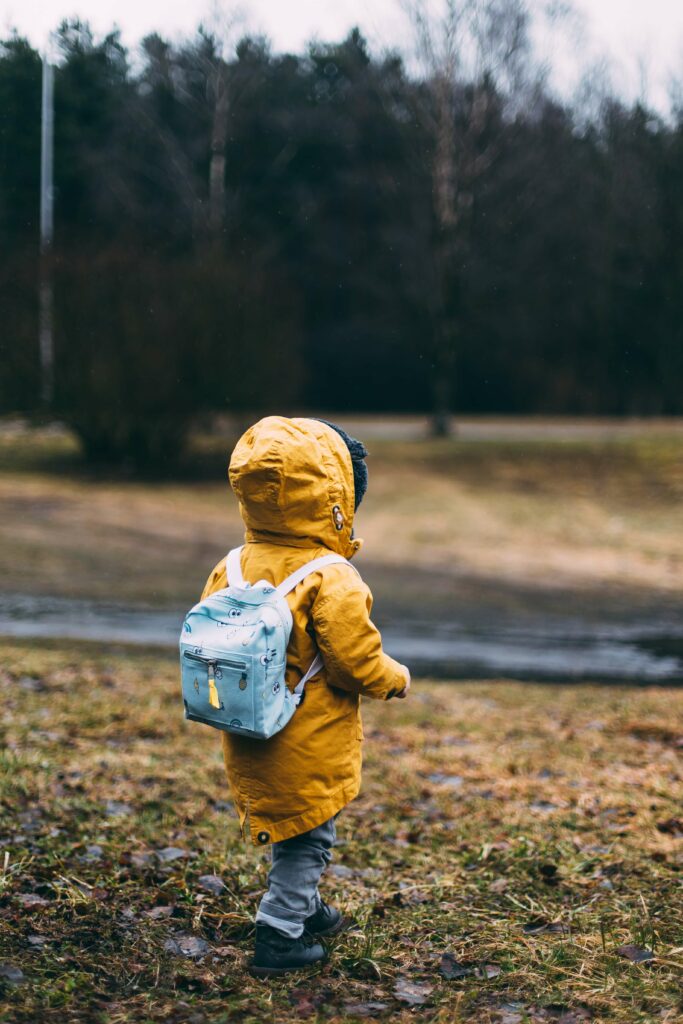 A young kid, walking outside in the bush with a yellow raincoat and a light blue backpack, exploring lessons and having a great time doing it.
