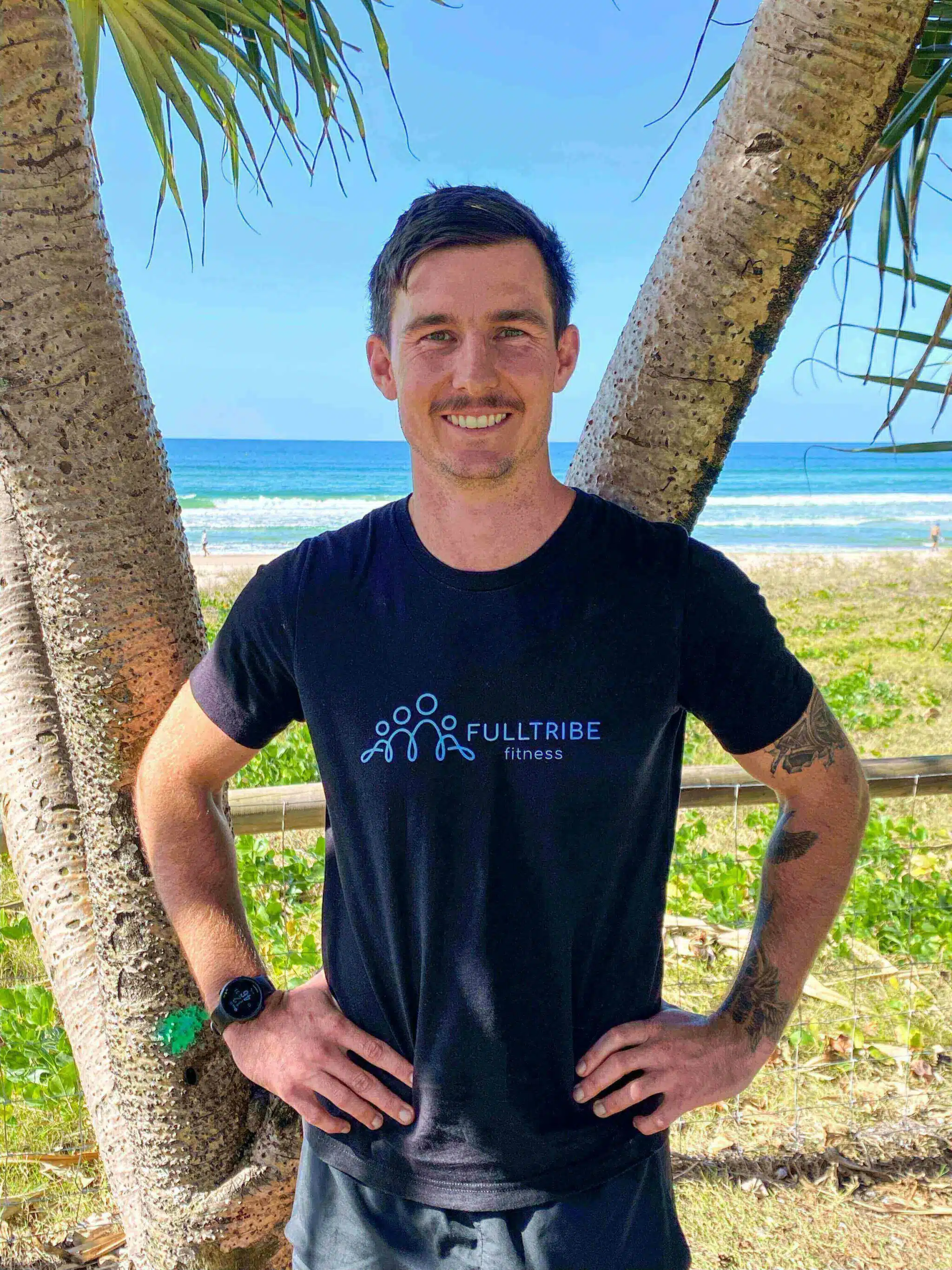 Jordan Pickard standing in front of a beach and palm trees. He has his hand on his hips and is wearing a black shirt with the Full Tribe Fitness logo on the front